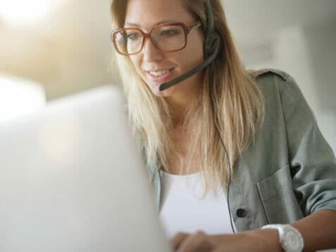 Eine blonde Frau mit Headset sitzt vor einem Laptop und arbeitet.