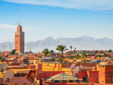 Stadtpanorama von Marrakesch mit Minarett vor Gebirgskette im Hintergrund unter blauem Himmel.