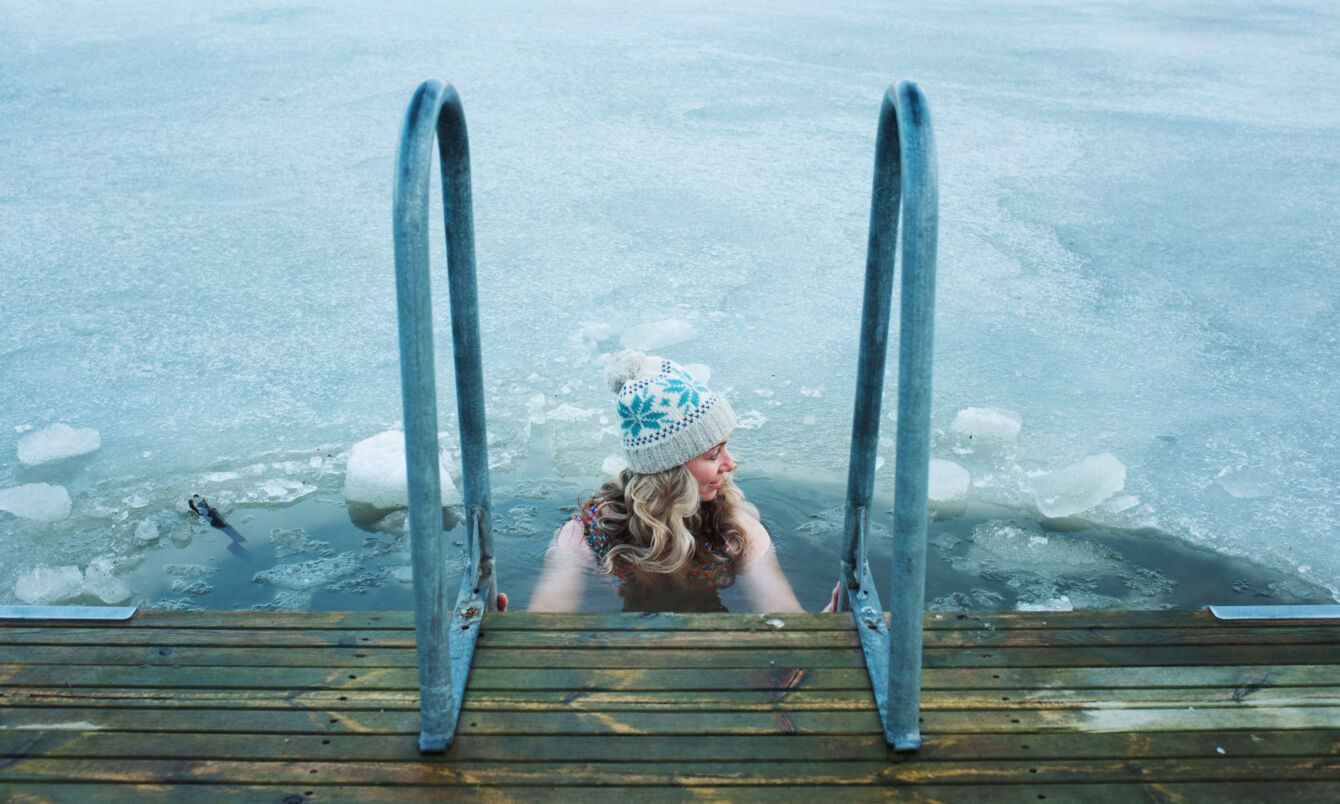 Eine Frau mit Wollmütze hält sich beim Baden in einem zugefrorenen See an dem Handlauf einer Treppe an einem Holzsteg fest.