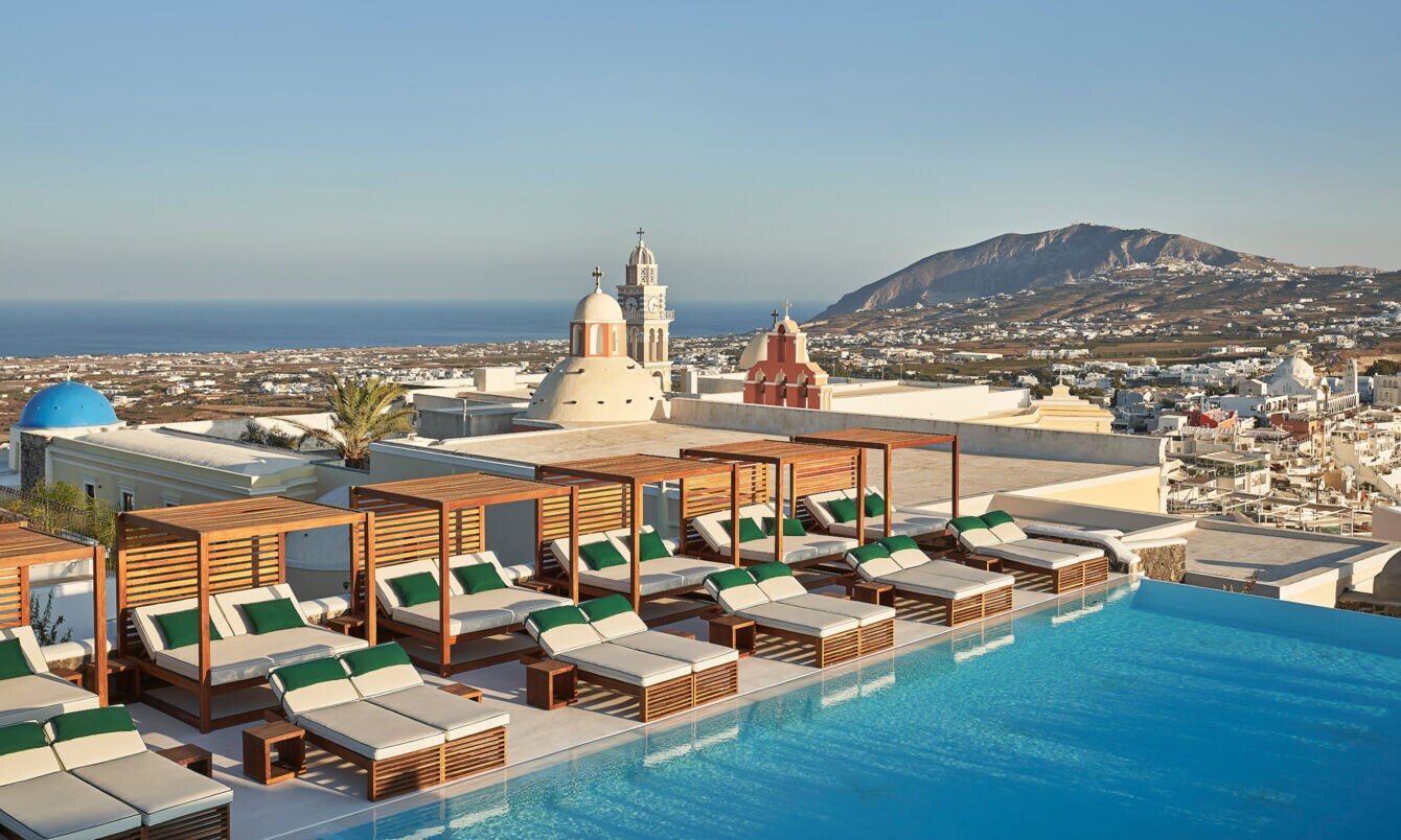 Dachterrasse mit zahlreichen Sonnenliegen am Infinity-Pool auf Santorini, im Hintergrund Meeresküste.