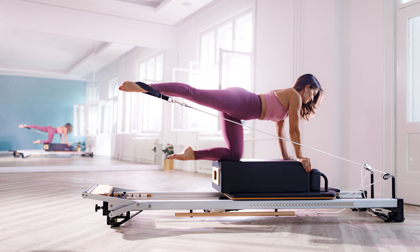 Eine Frau in pinker Sportkleidung kniet auf einer Box auf einem Pilates Reformer in einem hellen Raum.