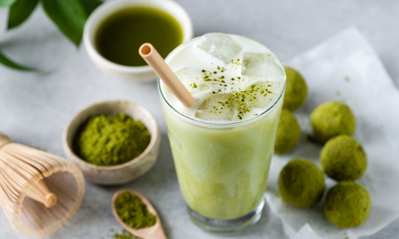 Ein Glas mit Matcha Latte und Strohhalm, daneben liegen grüne Kugeln, eine Schale mit grünem Pulver, ein Bambusbesen und eine Schale mit purem Matchatee.