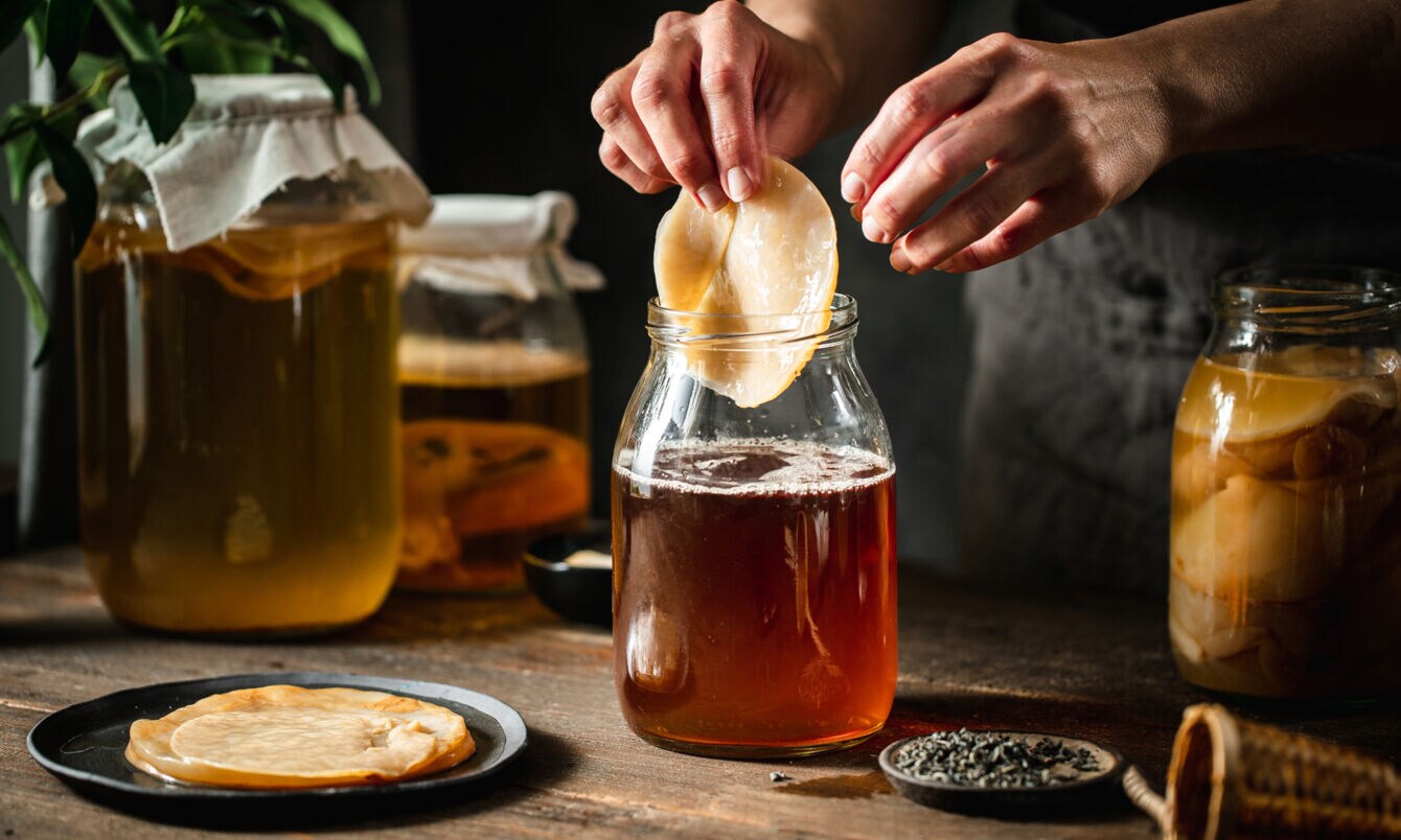 Ein Kombucha-Pilz wird von einer Hand in ein Gefäß mit brauner Flüssigkeit auf einem Holztisch gegeben, daneben weitere befüllte Einmachgläser.