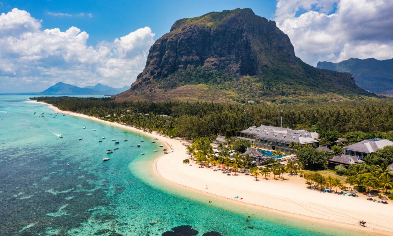 Küstenpanorama mit Hotelresort an einem palmengesäumten, weißen Sandstrand an türkisblauem Meer, im Hintergrund Berge.