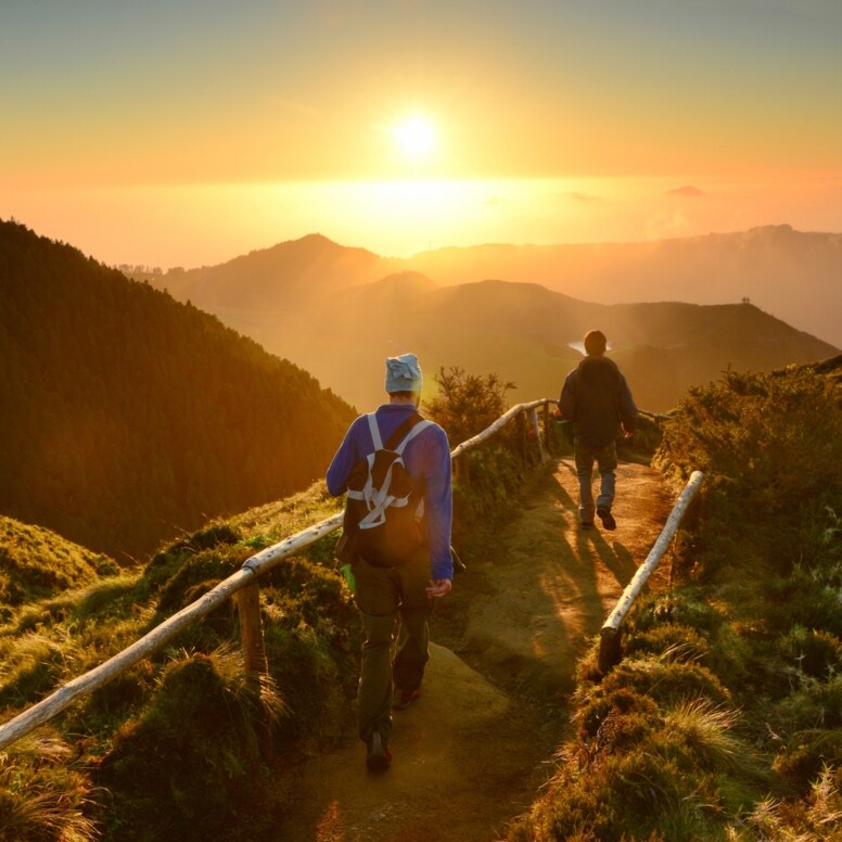 Rückansicht zweier gehender Personen auf einem Weg entlang eines Bergkamms inmitten einer grünen Gebirgslandschaft bei Sonnenuntergang.