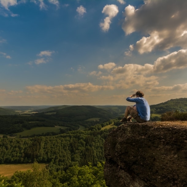 Ein Mann sitzt auf einem Felsen und blickt über die grüne Landschaft der Eifel