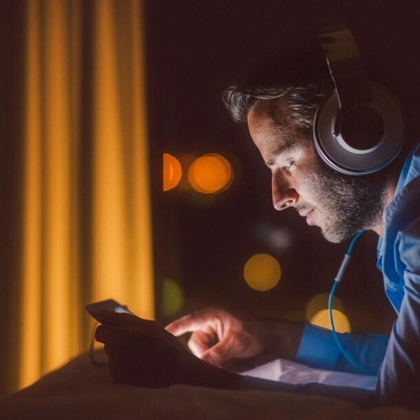 Ein Mann mit Kopfhörern und mobilem Gerät im Liegen, sein Gesicht erleuchtet vom Kunstlicht in der Dunkelheit