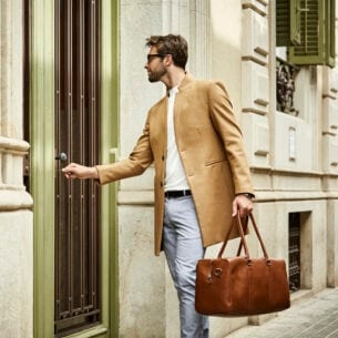Ein junger, moderner Mann mit Sonnenbrille, beigem Wollmantel und brauner Ledertasche schließt eine Haustür auf