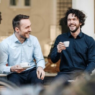 Zwei Männer genießen gut gelaunt einen Kaffee auf einer Bank vor einem Café