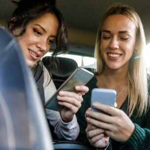 Zwei junge Frauen auf der Rückbank eines Autos schauen lächelnd auf ihre Smartphones