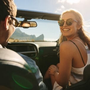 Eine junge Frau mit Sonnenbrille auf dem Beifahrersitz eines Cabriolets schaut lachend zu ihrem Partner hinüber, im Hintergrund eine Küstenlandschaft bei Sonnenschein.
