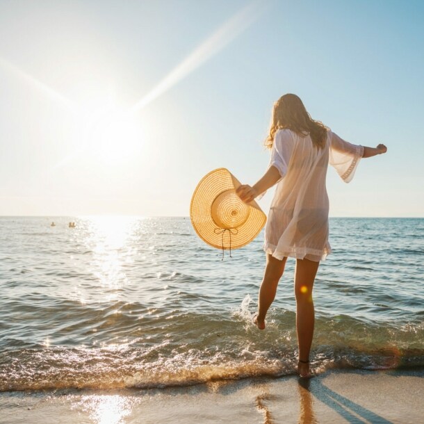 Eine junge Frau im weißen Sommerkleid und mit Strohhut in der Hand planscht mit ihrem Fuß im Wasser an einem Sandstrand am Meer bei Sonnenuntergang