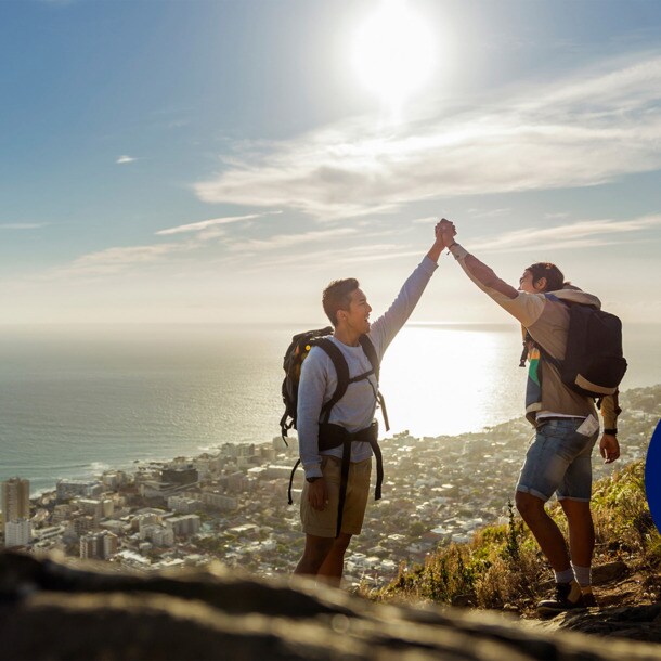 Zwei Wanderer mit Rucksäcken geben sich ein High Five auf einer Bergspitze, im Hintergrund eine Großstadt am Meer.