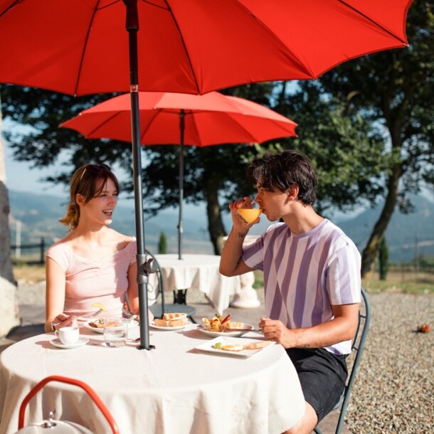 Ein Paar frühstückt auf einer Terrasse an einem Tisch bei Sonnenschein, im Hintergrund eine Hügellandschaft.