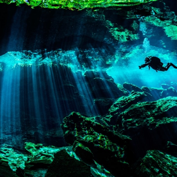 Eine Person in Taucherausrüstung taucht in einer von Wasser gefüllten Höhle. Lichtsäulen ergeben sich durch den starken Lichteinfall von oben im Wasser.