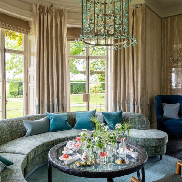 Luxuriöser Salon mit Kronleuchter und geschwungenem Sofa vor Panoramafenstern mit Blick in einen Garten.
