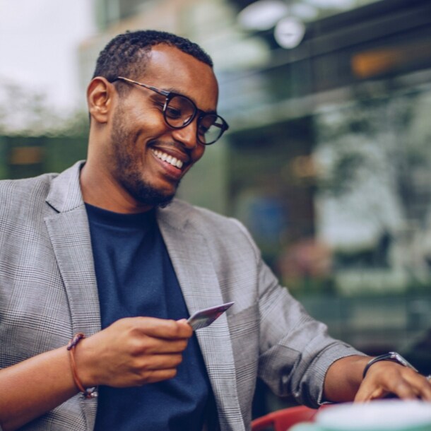 Ein junger, lächelnder Mann sitzt mit einer Kreditkarte in der Hand vor einem Laptop in einem Café.