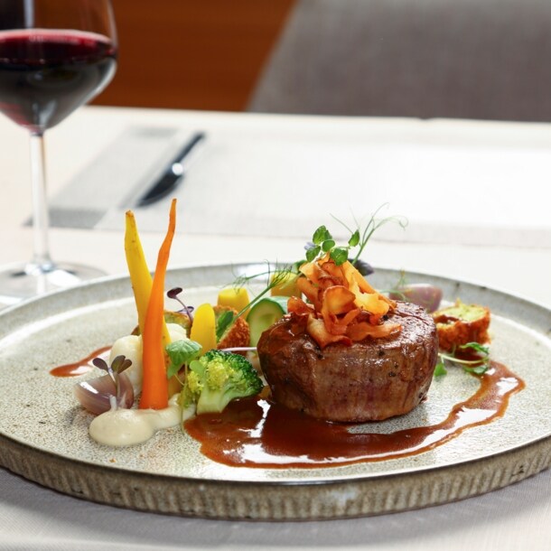 Ein Stück Steak an Gemüse, angerichtet auf einem Teller neben einem Glas Rotwein auf einem weiß gedeckten Tisch.