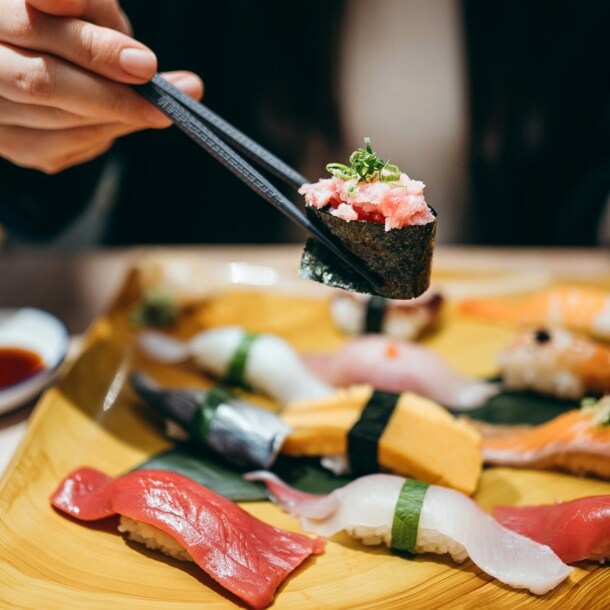 Eine Person hält eine Rolle Maki Sushi zwischen zwei Essstäbchen über einer Bambusplatte mit Nigiri auf einem Tisch in einem Restaurant.
