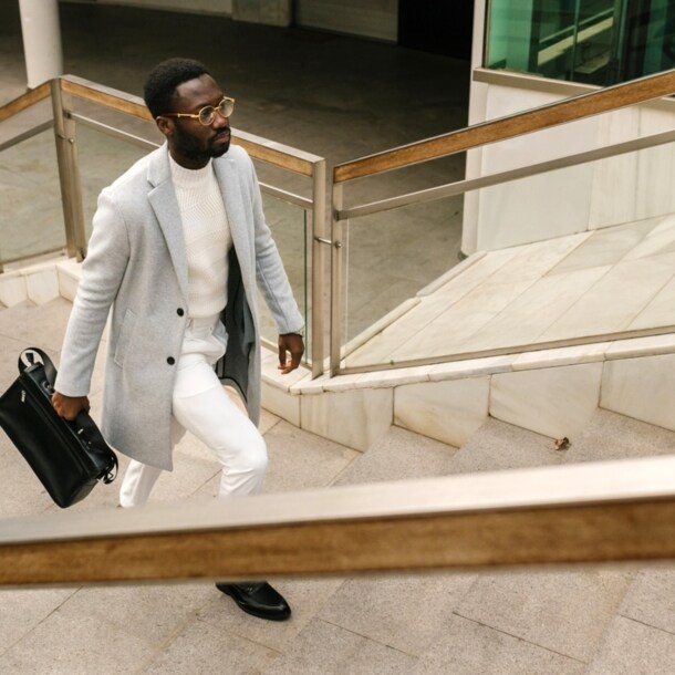 Ein elegant gekleideter Mann in einem hellen Outfit mit Wollmantel und schwarzer Aktentasche läuft eine Außentreppe hoch.