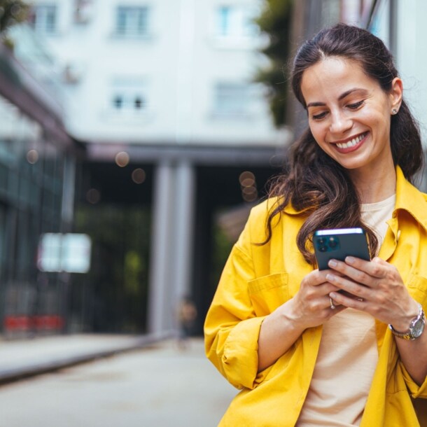 Eine Frau im gelben Hemd lehnt an einer Außenfassade und schaut lächelnd auf das Smartphone in ihren Händen.