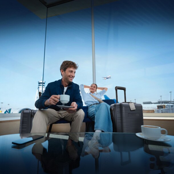 Zwei Personen sitzen nebeneinander in einer Flughafen-Lounge, neben sich jeweils einen Rollkoffer der Marke HORIZN, und trinken einen Kaffee.