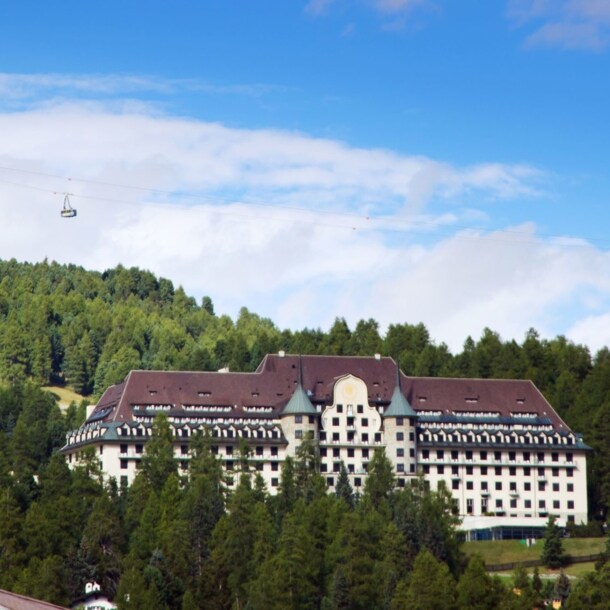 Ein Grandhotel mit zwei Türmen in einem Waldgebiet auf einem Berg, darüber fährt eine Seilbahn.