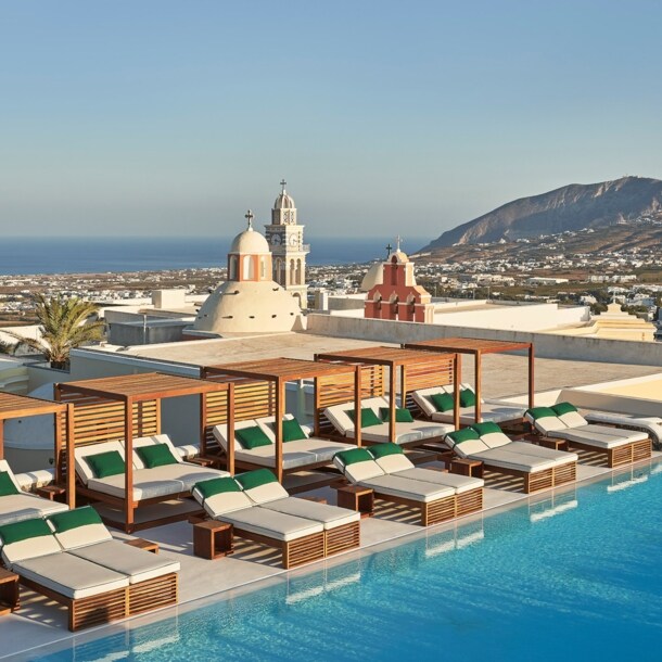 Dachterrasse mit zahlreichen Sonnenliegen am Infinity-Pool auf Santorini, im Hintergrund Meeresküste.