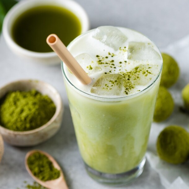 Ein Glas mit Matcha Latte und Strohhalm, daneben liegen grüne Kugeln, eine Schale mit grünem Pulver, ein Bambusbesen und eine Schale mit purem Matchatee.