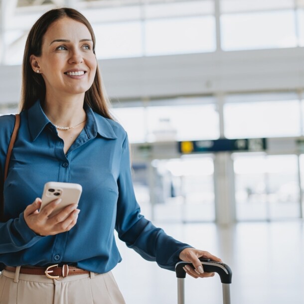 Eine Frau am Flughafen, die in einer Hand einen Koffergriff hält und in der anderen ein Smartphone.