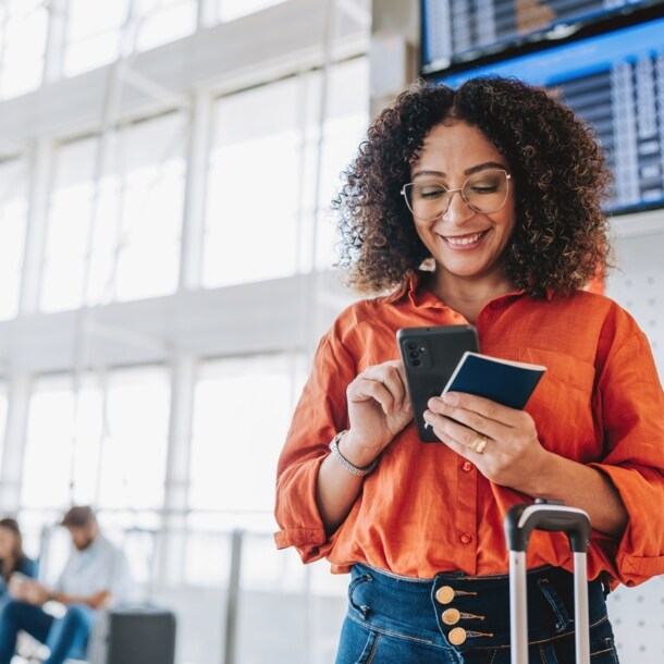 ine Frau bedient lächelnd ihr Smartphone in einem Flughafenterminal, in ihrer Hand ein Reisepass.