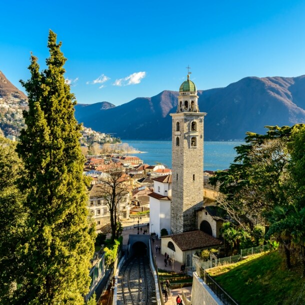 Stadtpanorama von Merano mit Zypressen und Glockenturm an einem See vor Bergpanorama.