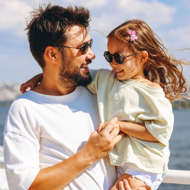 Ein lächelnder Mann mit Sonnbrille mit einem fröhlichen Mädchen mit Sonnenbrille auf dem Arm auf dem Außendeck eines Schiffes.