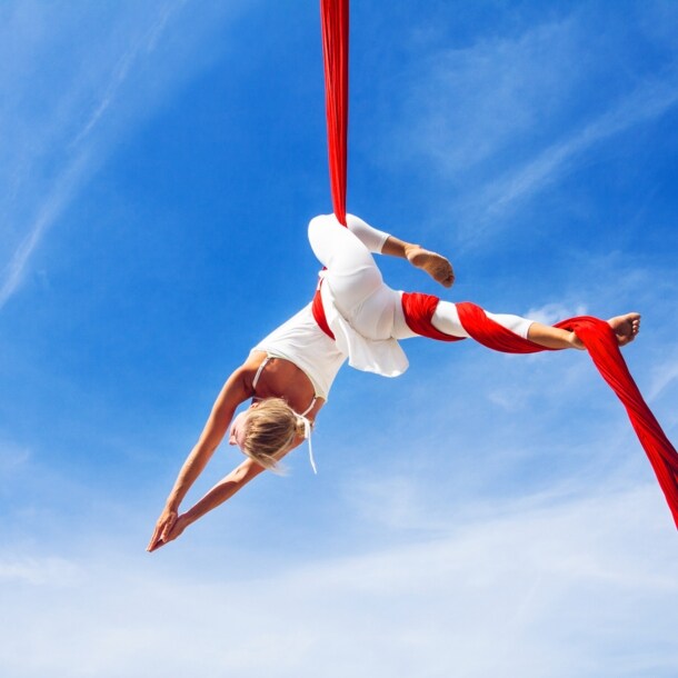 Untersicht einer Frau in weißer Kleidung in schwebender Yoga-Pose in einem roten Tuch vor blauem Himmel.