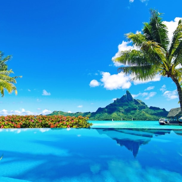 Von Palmen gesäumter Infinitypool an türkisblauer Lagune mit Blick auf eine Küste mit grüner Vegetation unter blauem Himmel.