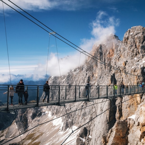 Personen auf einer Hängebrücke vor einer Felswand im Gebirge.