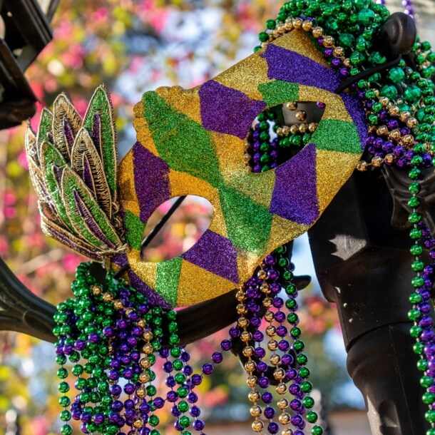 Eine Maske und Perlenketten in den Farben Grün, Gold und Violett an einer Straßenlaterne.