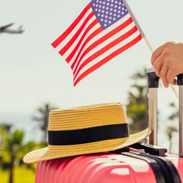 Ein Strohhut auf einem pinken Hartschalenkoffer, eine Hand am Griff hält eine kleine US-amerikanische Flagge, im Hintergrund Plamen und ein Flugzeug am Himmel.