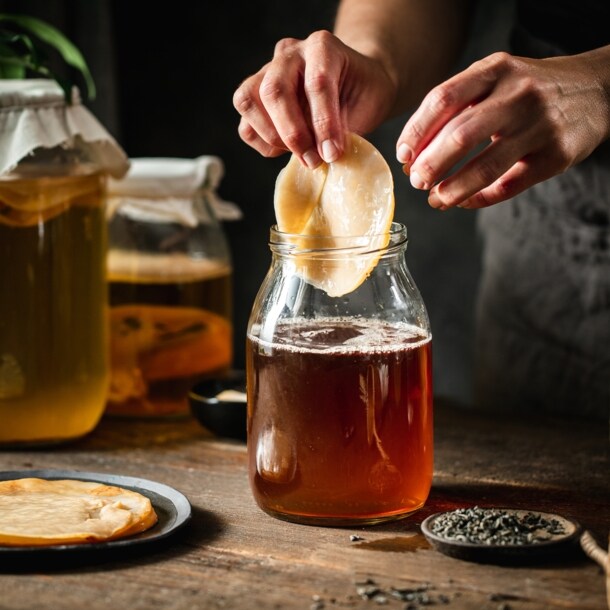 Ein Kombucha-Pilz wird von einer Hand in ein Gefäß mit brauner Flüssigkeit auf einem Holztisch gegeben, daneben weitere befüllte Einmachgläser.