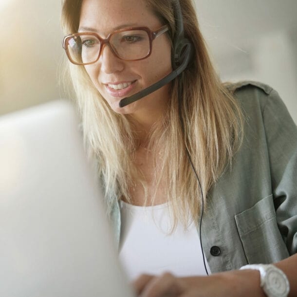 Eine blonde Frau mit Headset sitzt vor einem Laptop und arbeitet.