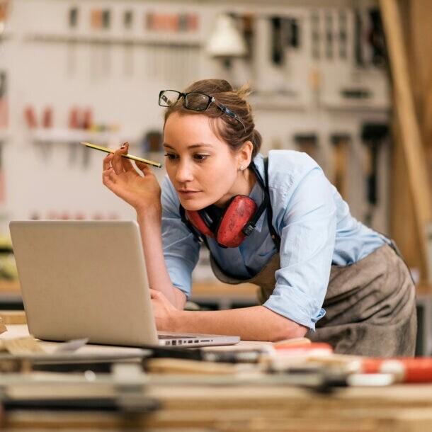 Foto einer Handwerkerin, wie sie an ihrer Werkbank steht und auf den vor ihr geöffneten Laptop blickt.