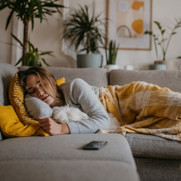 Eine Frau liegt schlafend mit einer Decke auf einer grauen Couch, im Arm hält sie einen Hund.