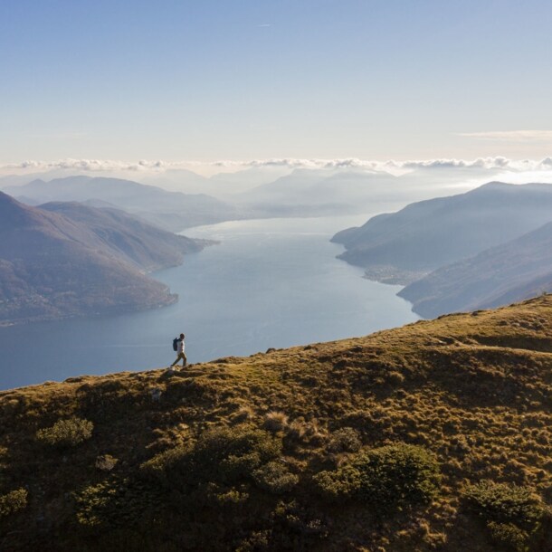 Panoramaaufnahme von einem Mann mit Rucksack auf einem Berg und dem Lago Maggiore im Hintergrund.