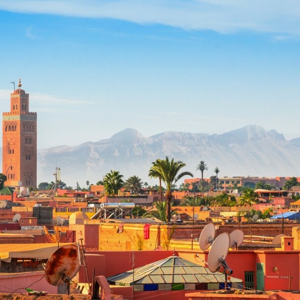 Stadtpanorama von Marrakesch mit Minarett vor Gebirgskette im Hintergrund unter blauem Himmel.