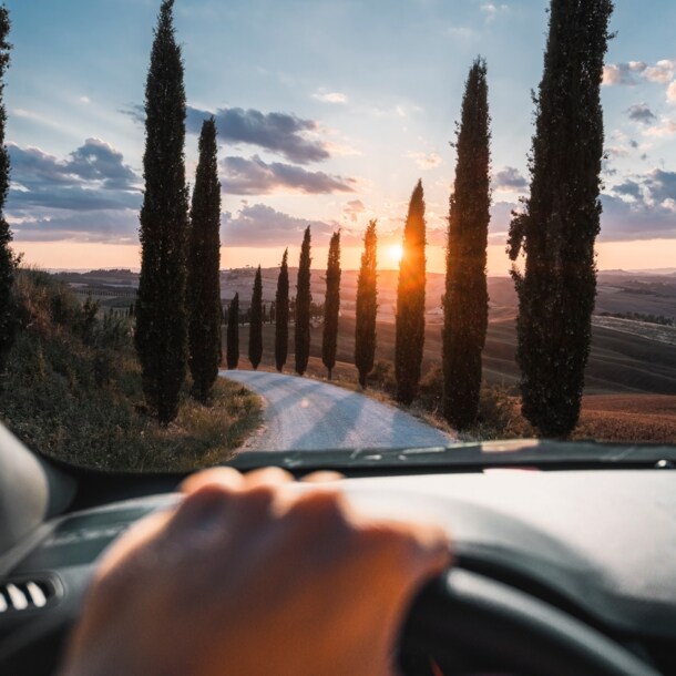 Blick auf eine mit Zypressen gesäumte Landstraße in Hügellandschaft bei Sonnenuntergang durch die Frontscheibe eines Autos, im Vordergrund eine Hand am Lenkrad.