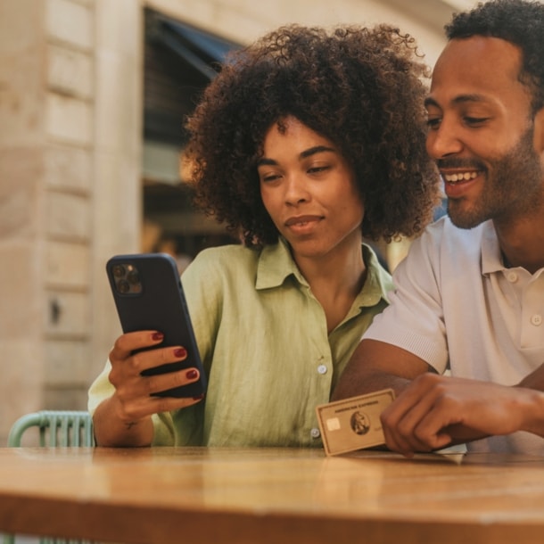 Ein Paar steht an einem Tisch auf einer Einkaufsstraße und schaut gemeinsam auf ein Smartphone, er hält eine goldene Kreditkarte von American Express in der Hand.