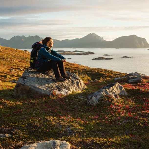 Eine Frau mit Wanderausrüstung, die bei Sonnenuntergang auf einem großen Stein sitzt und aufs Wasser blickt, im Hintergrund Berge.