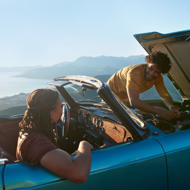 Ein Paar in einem liegengebliebenen Cabriolet an einer Meeresküste bei Sonnenschein, er kontrolliert den Motor, sie sitzt auf dem Beifahrersitz.