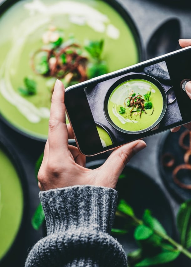 Zwei Hände halten Smartphone, das eine grüne Suppe in Schüssel fotografiert