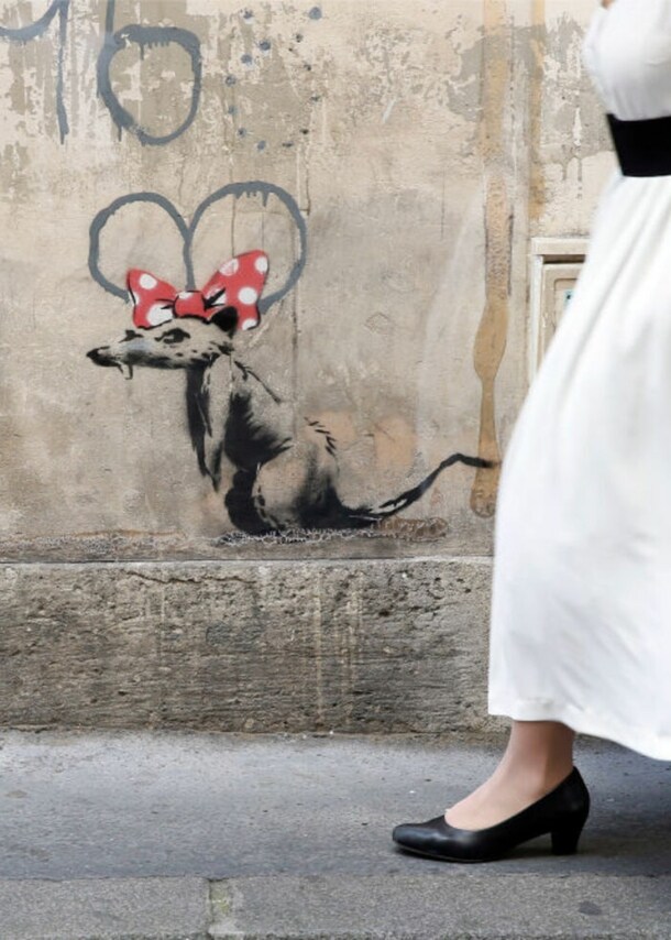 Eine Frau in langem, weißen Kleid läuft an einem Banksy-Werk vorbei: eine Ratte mit roter Minnie-Mouse-Schleife auf dem Kopf.